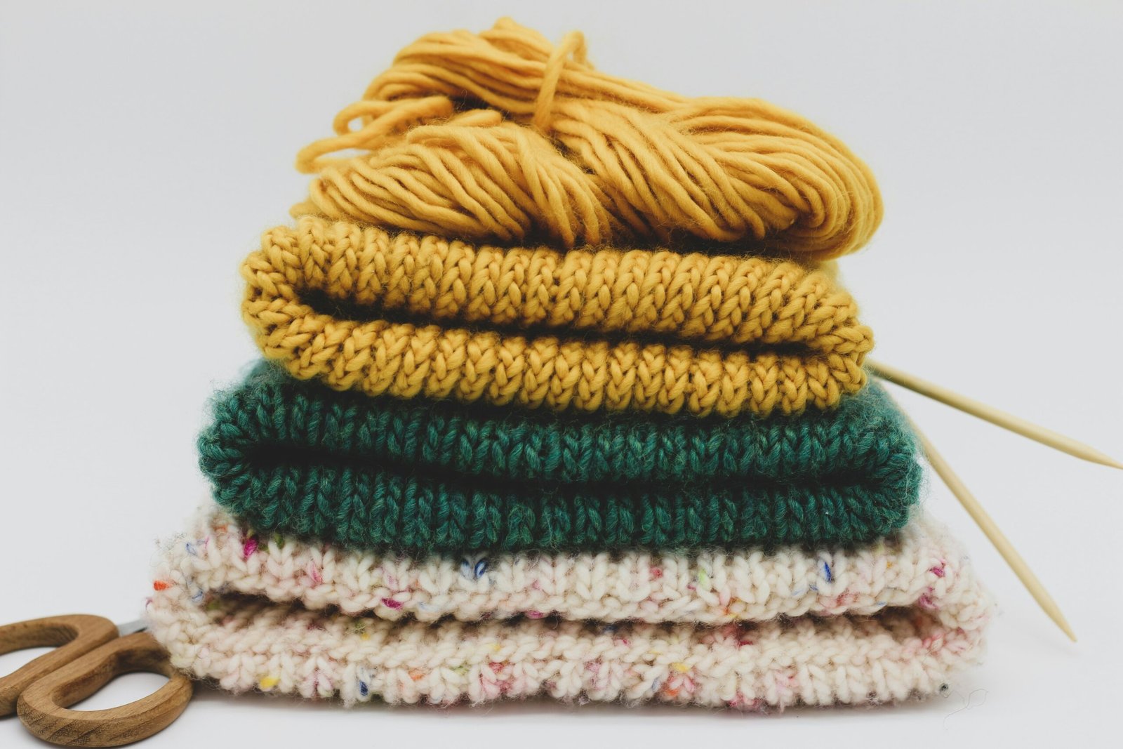 A neatly arranged vertical stack of three folded, hand-knitted beanies in mustard yellow, forest green, and cream-speckled yarn. On top sits a loose bundle of matching yellow yarn, while a pair of wooden-handled scissors and two light-colored wooden knitting needles rest beside the pile on a clean white surface.