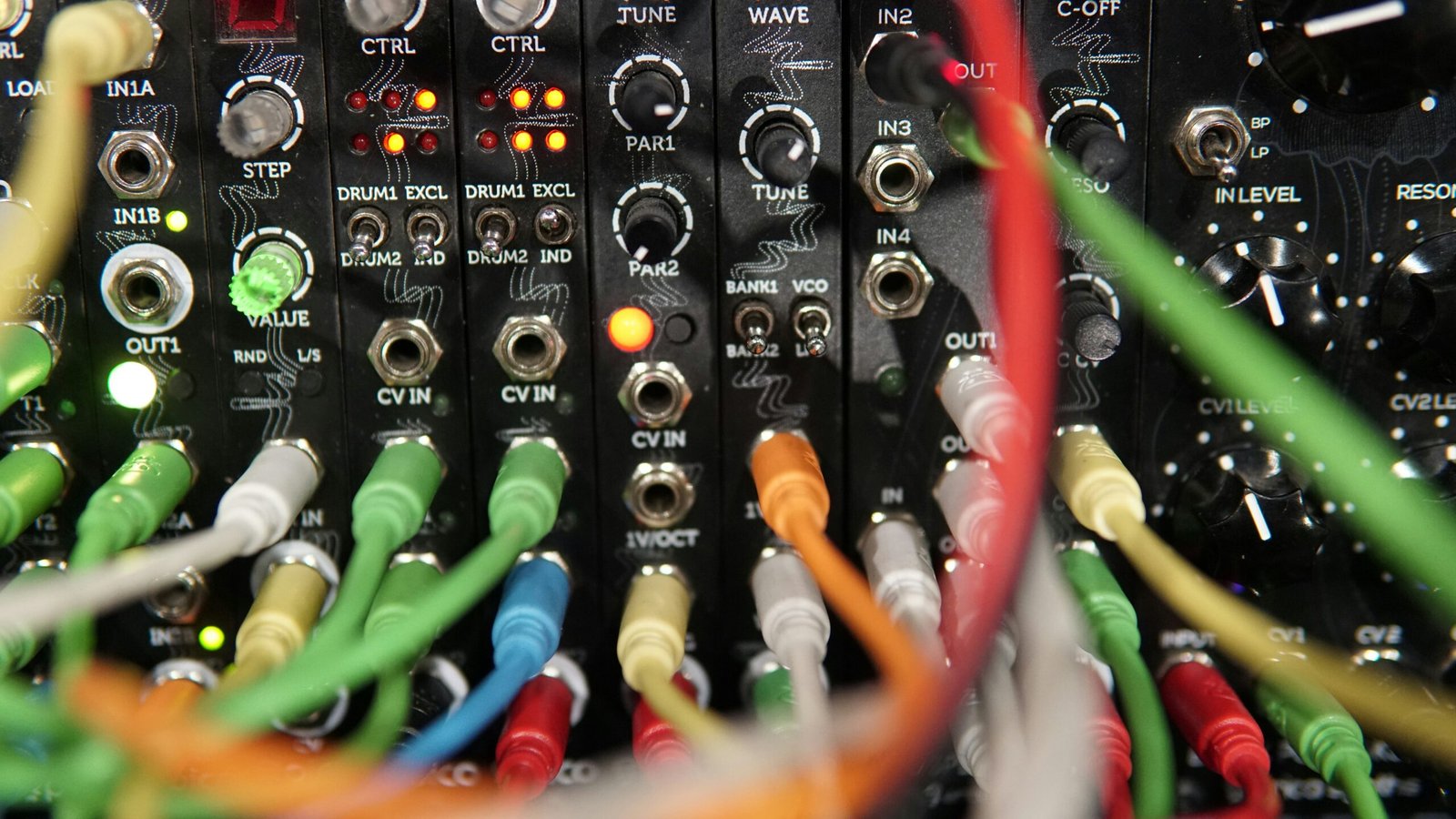 A dense, close-up view of a black Eurorack modular synthesizer faceplate, specifically the Erica Synths Pico System 1, showing multiple thin modules with various knobs, switches, and illuminated LEDs. A chaotic web of colorful patch cables in red, yellow, green, blue, and white crisscrosses the frame, plugged into numerous 3.5mm input and output jacks to create a complex audio patch.