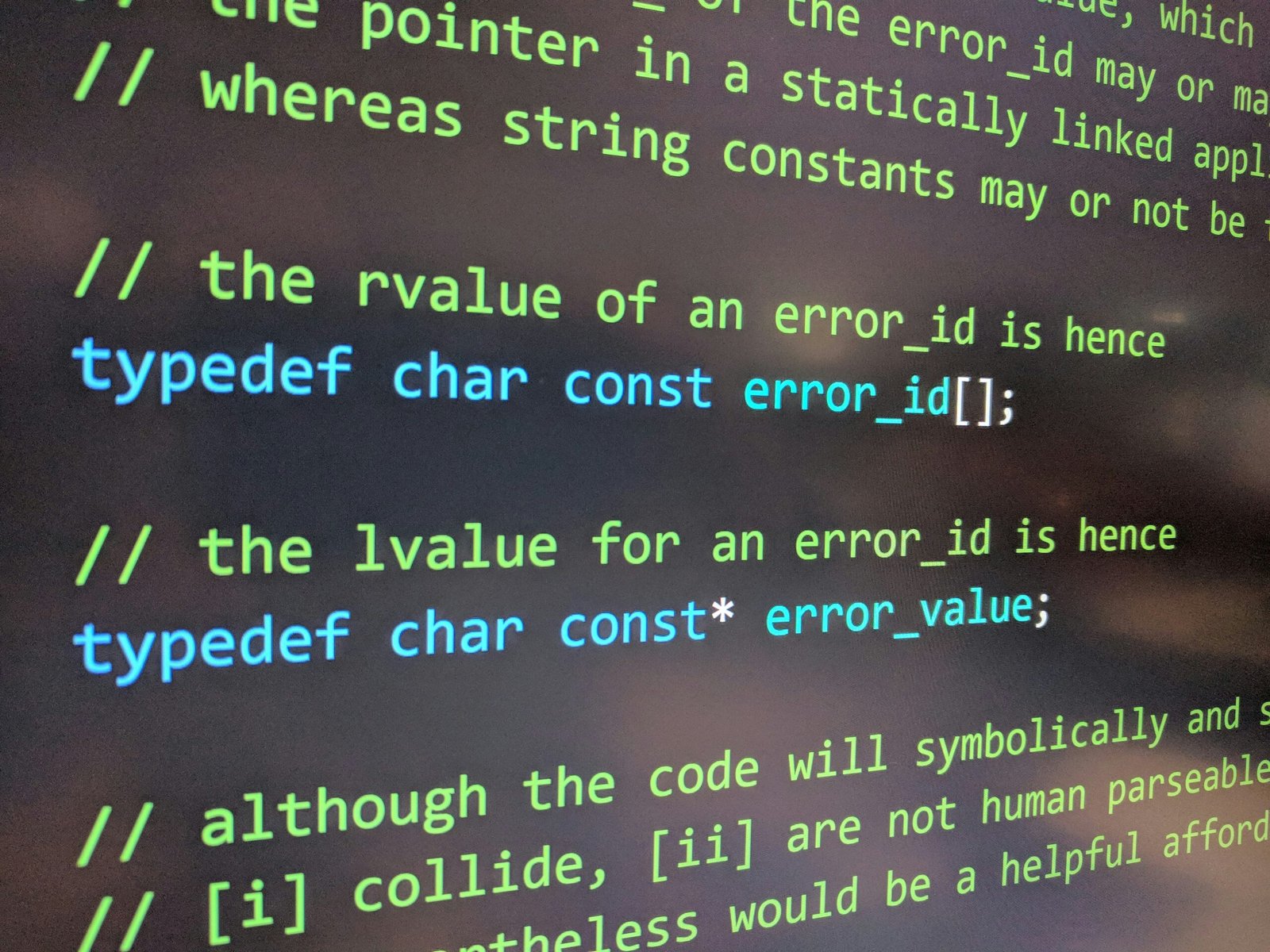 A close-up, slightly angled photo of a computer screen displaying C or C++ source code on a dark background. The text features green comments explaining the logic behind "rvalue" and "lvalue" for error identification, alongside two teal-colored typedef statements defining error_id as a constant character array and error_value as a constant character pointer.
