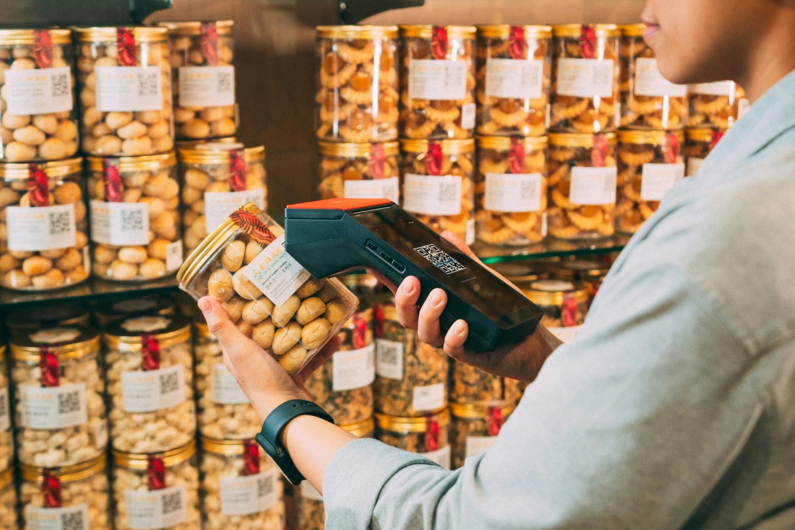 Close-up of a person in a light blue shirt using a handheld digital scanner to scan a QR code on a jar of cookies in a well-stocked retail store.