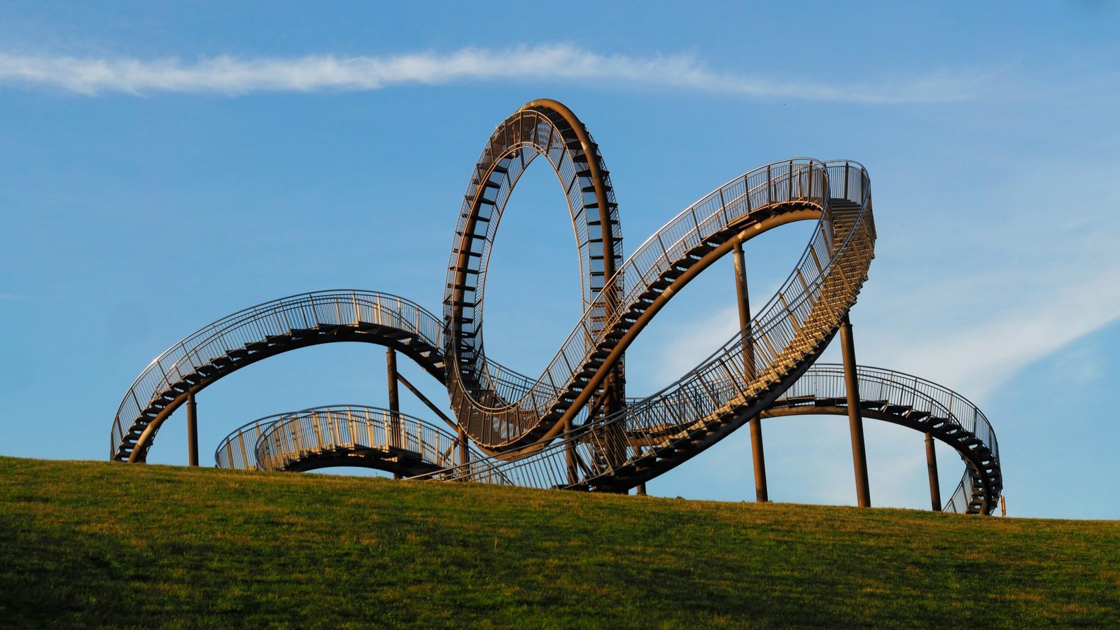 A large, silver steel sculpture resembling a winding roller coaster with a vertical loop sits atop a grassy green hill under a clear blue sky. The structure features walkable stairs and handrails, with the sun casting a warm, golden light across its metallic surface.
