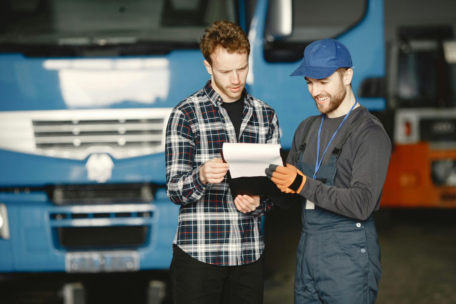 Truck Mechanic and Customer Reviewing Maintenance Checklist A man in a plaid shirt and a mechanic in blue overalls and a cap stand together in a garage, reviewing a document on a clipboard in front of a large blue truck.