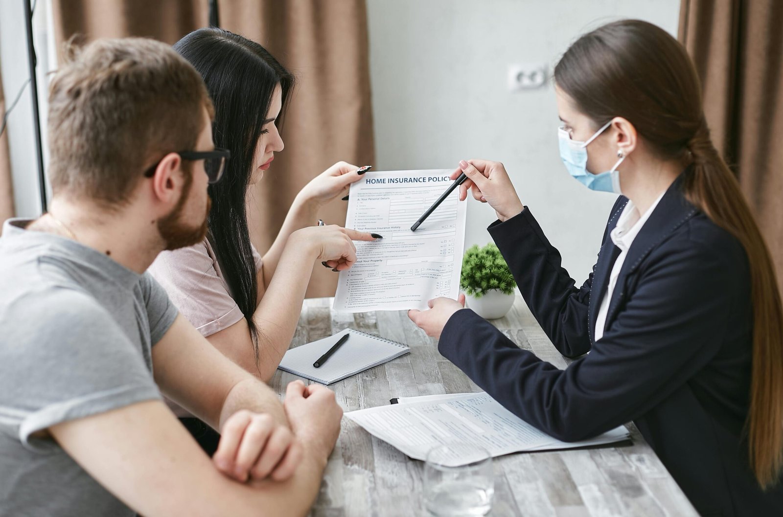 Young Couple Reviewing Home Insurance Policy with Agent An insurance agent wearing a blue surgical mask and a navy blazer points to a "Home Insurance Policy" document with a pen while a young couple reviews it across a wooden desk in an office setting.