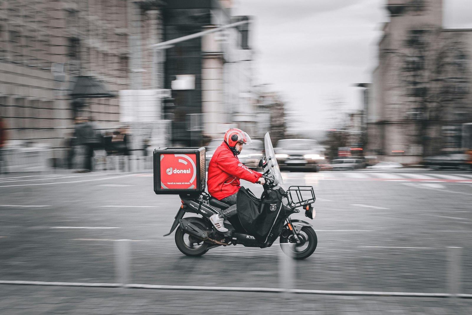 "A delivery rider in a red jacket and helmet riding a black scooter with a red 'donesi' delivery box through a busy, blurred city square.