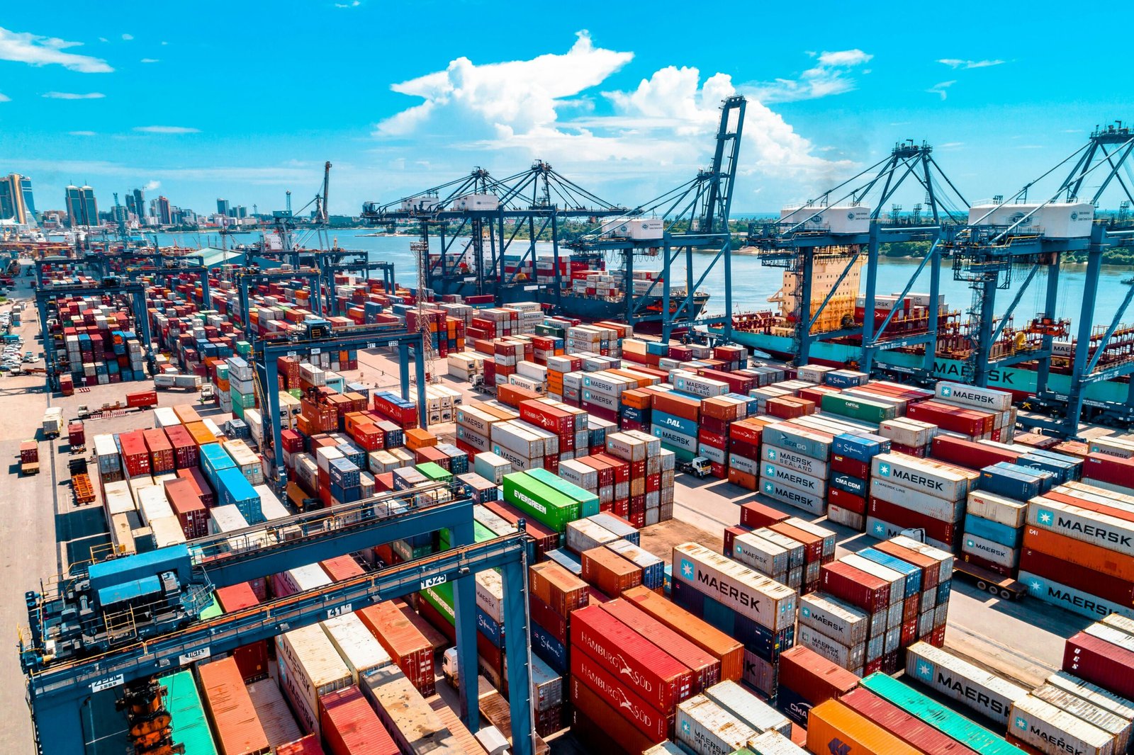 Aerial view of a busy shipping terminal with rows of colorful cargo containers, massive blue gantry cranes, and large container ships docked at the waterfront.