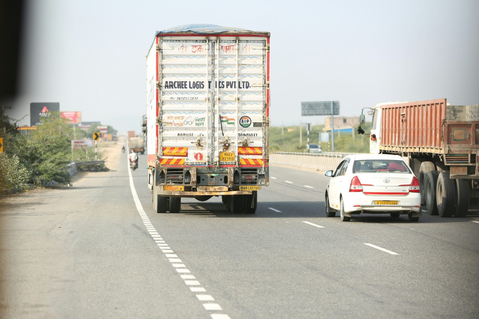 arjun-baroi-ZqvYqnlkicw-unsplash A rear view of a white commercial truck from Archee Logistics Pvt Ltd. driving on a multi-lane paved highway in India, alongside a white sedan and a large brown flatbed truck under a bright, clear sky.