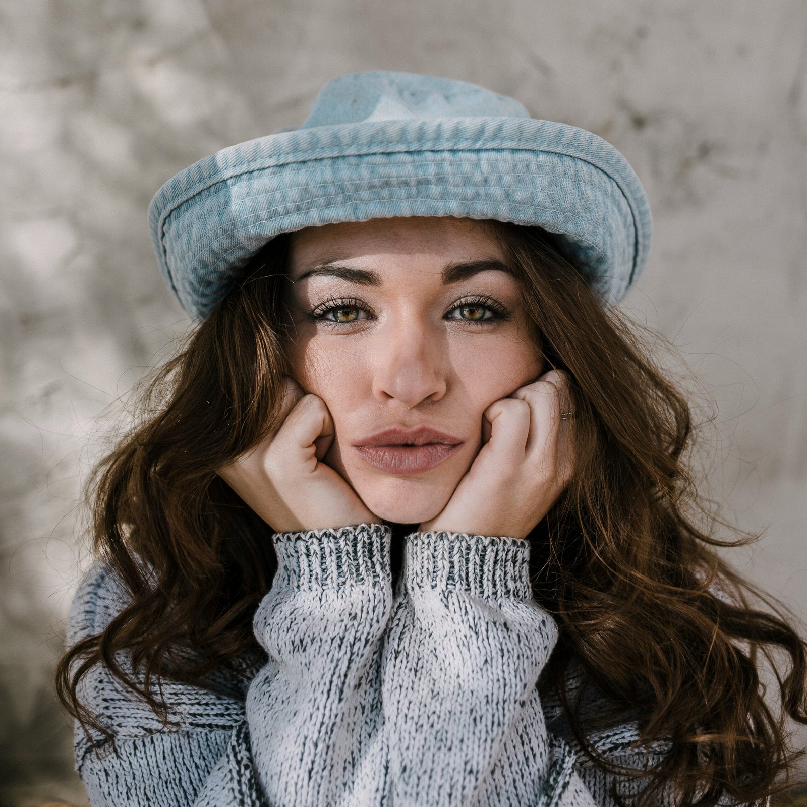 Close-up portrait of a woman wearing a hat and sweater, resting her face on her hands and looking directly at the camera with a calm expression.
