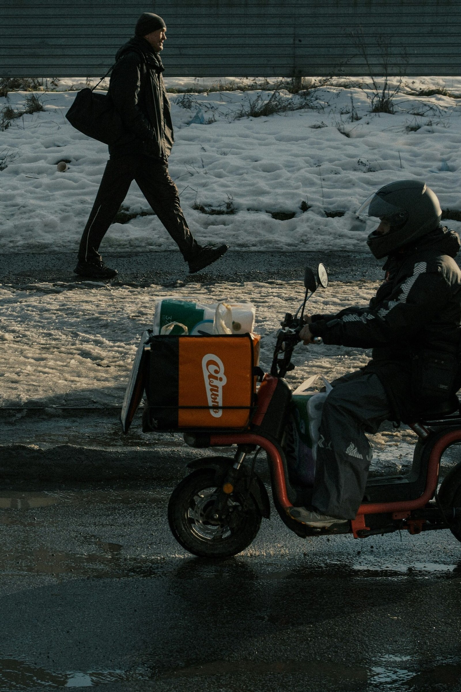 A delivery rider on an orange electric scooter with a large cargo box in the foreground, while a man in dark winter clothing walks along a snowy roadside in the background.
